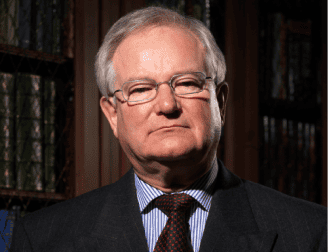 An older man wearing glasses, a suit, and a tie stands in front of bookshelves.
